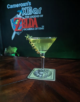 An emerald green drink in a martini glass is garnished with a spiraled lime peel with a sprig of thyme threaded through it. Behind the bar the chalkboard glows a dull green light and has "Cameroxn's XBar The Legend of Zelda Ocarina of Time" written on it.