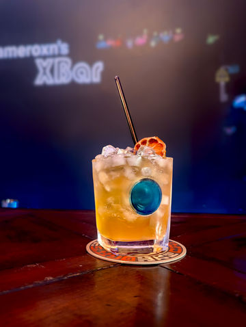 A short glass filled with pebble ice and an amber-brown liquid, garnished with a dehydrated citrus wheel and a black straw. A round blue accent is embedded on the front of the glass, evoking Claptrap’s eye.