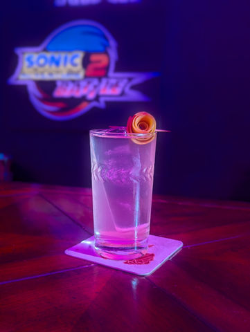 A bright pink drink stands in a tall glass with a wrapped grapefruit rose skewered on top. The board behind the drink says Sonic Adventure 2 Battle