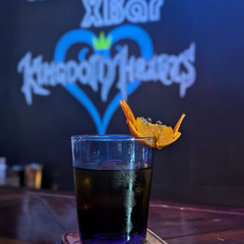 A dark orange drink in a blue-tinted glass. The glassware has a small crab garnish made out of a clementine and clove sitting on the edge with it's orange-skin claws in the air.