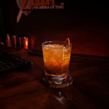 An orange and brown drink in a glass with ice on a bartop. A dark piece of dried citrus garnishes the glass. The dark red chalkboard behind says "Zelda"