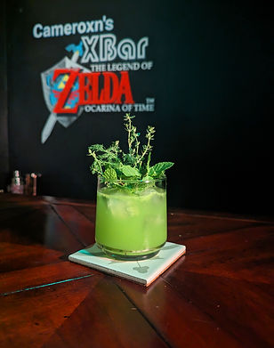 A clouded green cocktail garnished lavishly with various herbs including mint, thyme, and dill. The chalkboard behind says "Cameroxn's XBar The Legend of Zelda Ocarina of Time"