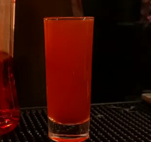 A dark red shot in a tall shooter glass. It stands upon a black rubber bar mat.