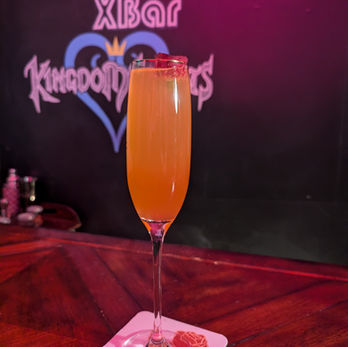 A tall orange-tinted drink in a champagne flute. A small rosebud floats on top of the drink.