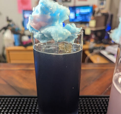 A dark purple-blue liquid in a highball glass. There is a blue tuft of cotton candy sitting atop the glass, seemingly floating.