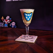 A yellow drink with a white foam head sits in a stemmed glass adorned with a blue and white shield.