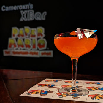 An orange cocktail in a coupe glass perched upon a bartop. Atop the glass rests a small paper airplane. The glass stands on a sheet of paper adorned with images of Paper Mario. A blackboard behind says "Cameroxn's XBar Paper Mario The Thousand-Year Door"