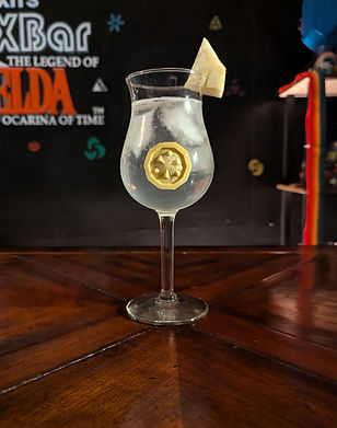 A clear drink in a tall curved glass with ice. The glass has a pineapple wedge hanging on the rim and a yellow emblem on it depicting three dots and triangles.