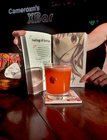 A vibrant reddish-orange cocktail sits centered in a short rocks glass on a floral coaster. Behind it is an open manga volume of Fruits Basket, showing the title "Feelings of Gratitude" and a close-up of the character Tohru Honda. A small smear of pickled plum (umeboshi) is visible on the back of the glass. Hands are gently holding the book open, with a warm spotlight on the drink and pages.