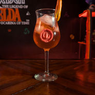 A red drink in a tall curved glass. The glass has one red-orange pepper hanging on the rim and an emblem depicting a flame adorns the front.