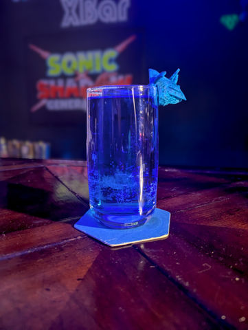 A curved tall glass filled with a royal blue beverage with noticeable bubbles sits on a blue hexagonal coaster. A blue spiked garnish sits on the side of the glass. The board behind says "Sonic x Shadow Generations".