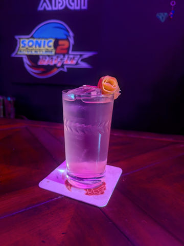 A tall pink drink stands in a florally adorned glass. Atop the drink is a small curled rose made from grapefruit peel. The board behind the drink says Sonic Adventure 2 Battle.