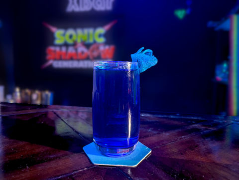 A curved tall glass filled with a royal blue beverage with noticeable bubbles sits on a blue hexagonal coaster. A blue spiked garnish sits on the side of the glass. The board behind says "Sonic x Shadow Generations".