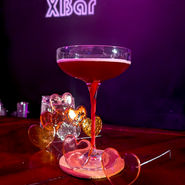 A coupe glass filled with a slightly brighter red mocktail with a noticeable white foam layer, also set on a coaster with a magenta ribbon winding up the stem. Rose-tinted candy heart spectacles rest in front of the glass, and a collection of multicolored isomalt hearts sits behind it. The Cameroxn’s XBar logo glows on the purple backdrop, tying the presentation together.