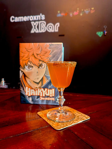 A bright orange cocktail called “Fly!! (Karasuno High)” sits in an ornate stemmed glass with a golden sugar rim, placed on a patterned coaster atop a dark wooden bar. Behind it stands the Haikyu!! anime season 3 box set featuring Shoyo Hinata with intense orange hair and a determined expression. The blurred background shows the text “Cameroxn’s XBar.”