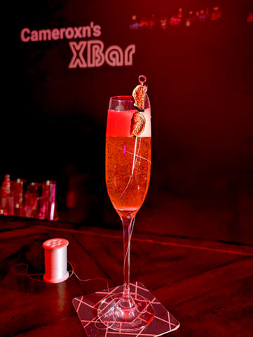 A tall flute glass filled with a deep red sparkling drink, topped with noticeable layer of white foam and rising bubbles. The garnish is two skewered dried fig halves on a needle-like pick with a circular top, with a white thread wrapped across the glass and leading to a spool placed on the table, echoing Hornet’s weapon and silk threads.