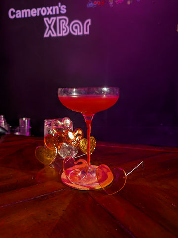 A coupe glass filled with a slightly brighter red mocktail with a noticeable white foam layer, also set on a coaster with a magenta ribbon winding up the stem. Rose-tinted candy heart spectacles rest in front of the glass, and a collection of multicolored isomalt hearts sits behind it. The Cameroxn’s XBar logo glows on the purple backdrop, tying the presentation together.