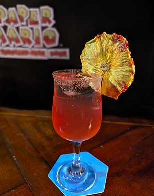 A red drink in a tapered and stemmed glass sits on a blue hexagon coaster. The drink's red hue has a gradient that fades from a transparent soda to a deep pink gin. The drink sports a dried pineapple wedge on its rim, reminiscent of a flower.