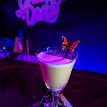 An opaque drink in a triangular coupe is adorned with a half-crescent of blue petals. An orange butterfly seems to hang on the side above the petals. The drink has two distinct layers, white with a purple light on top and yellow on the bottom. The text "Dream Daddy" is visible on the backdrop.