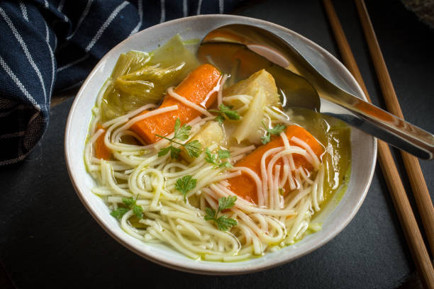 Bowl of noodle soup with carrots, greens, and a sprig of cilantro. Set on a dark table with chopsticks and a striped napkin.