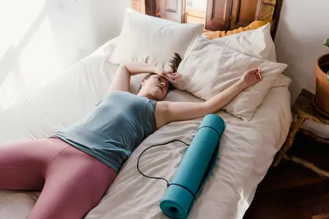 Woman in workout clothes rests on a bed with a blue yoga mat beside her, looking tired. Bright natural light in a cozy room.