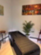 A massage bed in a warmly lit room with a plant, stool, and paintings on the walls. The colorful art depicts an autumn scene.