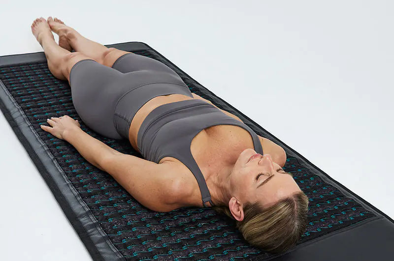 Woman in gray activewear lying on a black PEMF mat with turquoise details. She's relaxed, eyes closed, in a minimalistic bright setting.