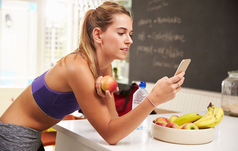 Woman in workout gear holds apple and phone in kitchen. Bowl of fruit and water bottle nearby. Blackboard with writing in background.