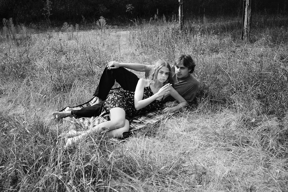 A couple relaxes on a blanket in a grassy field, girl in a floral dress, black and white photo, calm and serene atmosphere. "Happy Springs" by Patrick Bienert