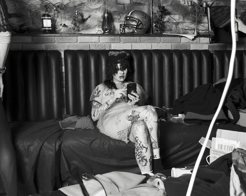 Tattooed person with dark hair sits on a cluttered room's couch, focused on their phone. "Wet ground" by Aria Shahrokhshahi
