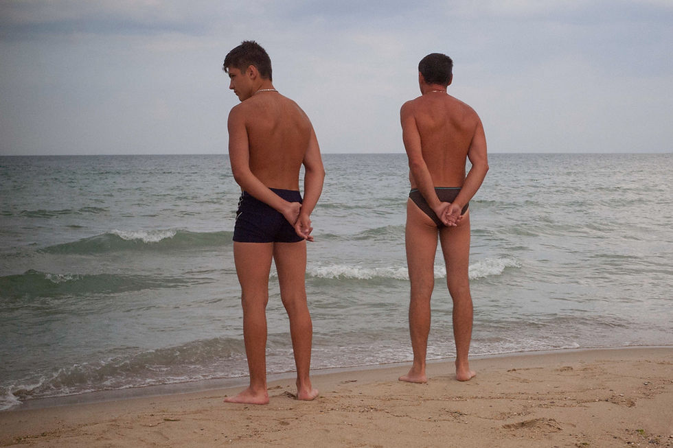 Two people in swimsuits stand on a sandy beach, facing the sea with hands behind their backs. "Happy Springs" by Patrick Bienert