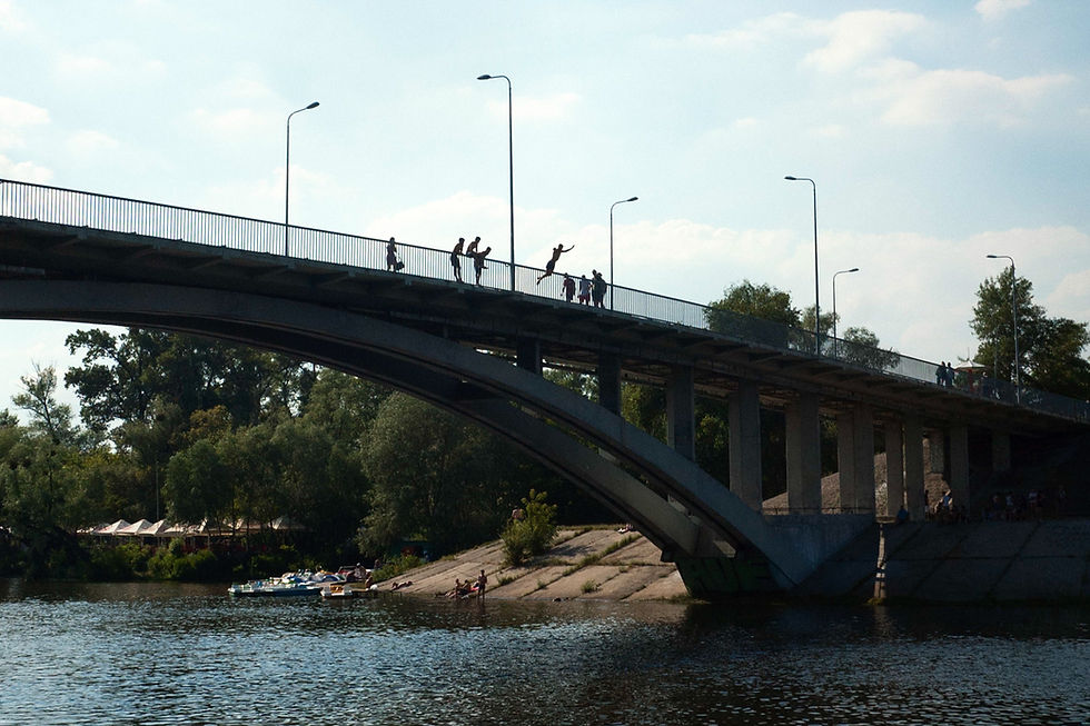 People jump off a bridge into a river, surrounded by trees under a clear sky in Kyiv. "Happy Springs" by Patrick Bienert