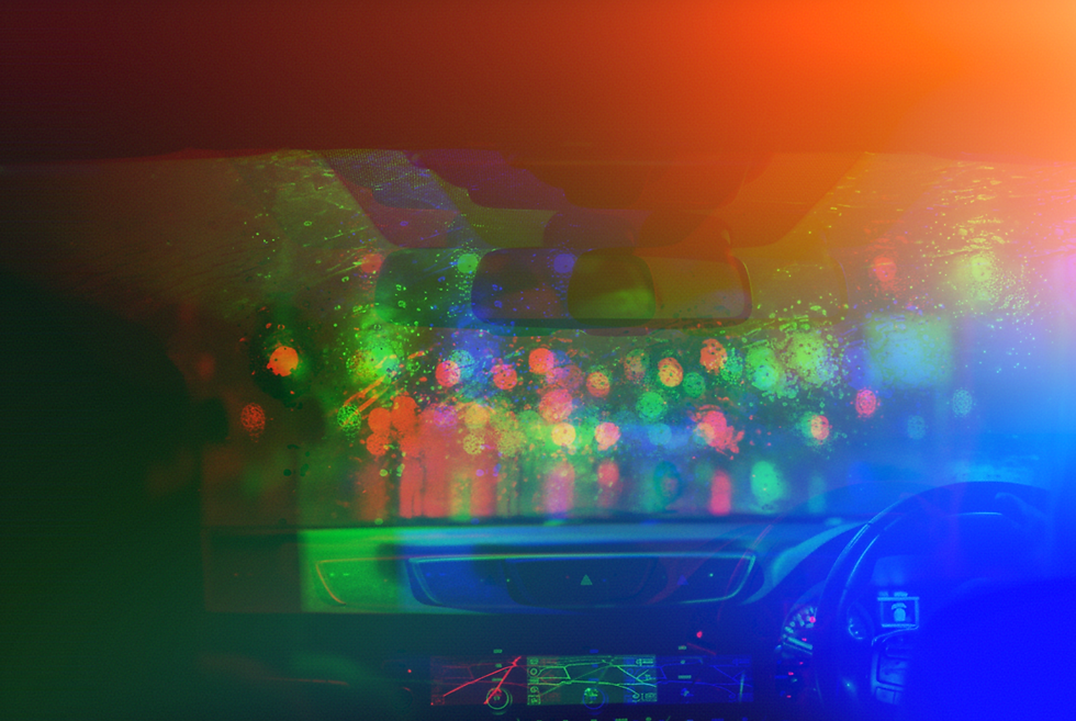View from a car's interior at night through a rain-streaked windshield, showing the dashboard and blurred blue and orange str