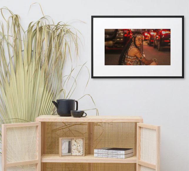 Woman with dreadlocks, face paint, and a patterned outfit sits on a street with cars and red brake lights in the background.