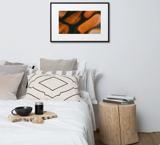 Abstract aerial landscape print (orange, white, and blue) hanging above a modern bed with neutral pillows and a wooden bedsid