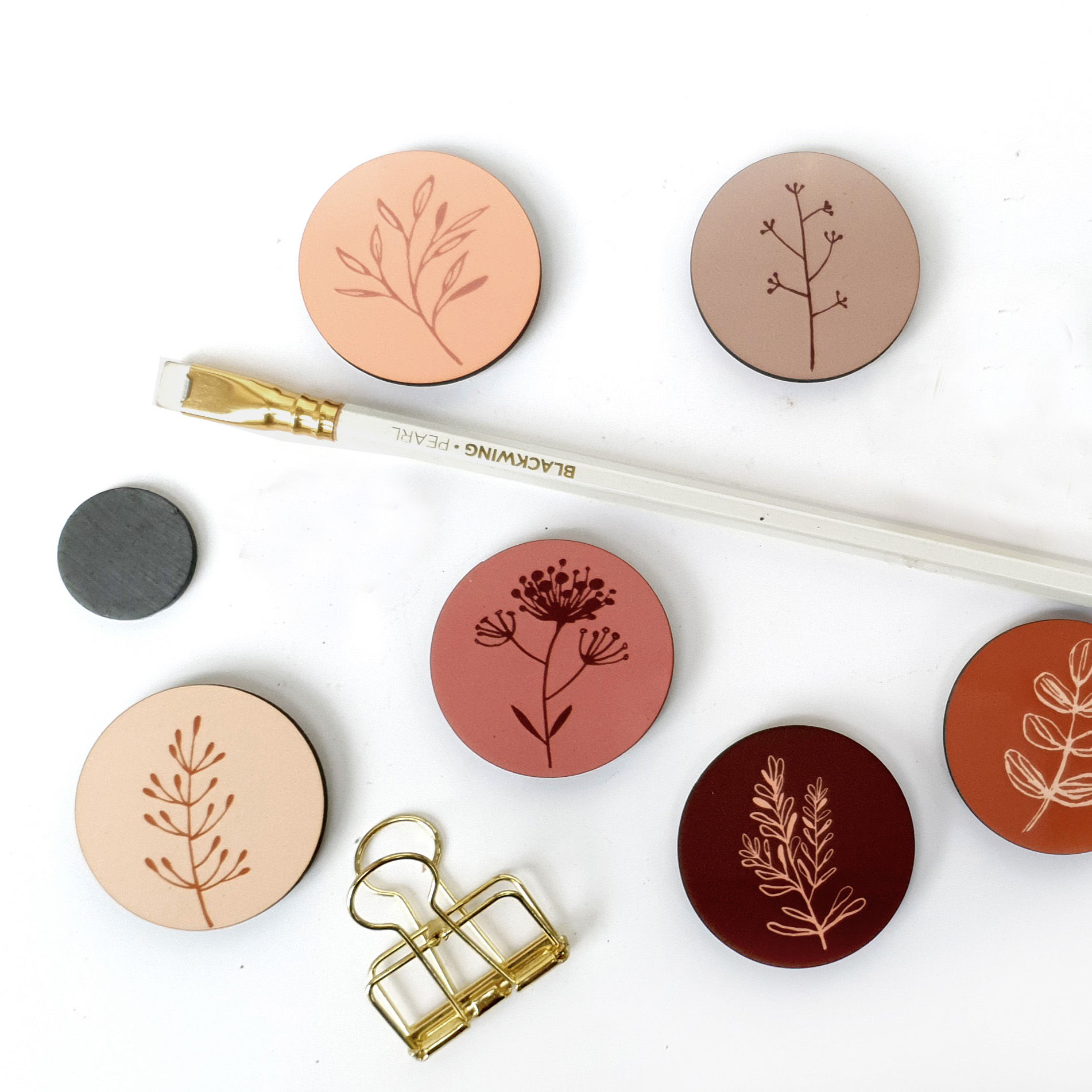 מגנטים עגולים עם איורים בוטניים, פרושים על שולחן ליד מכחול ועיפרון זהוב.