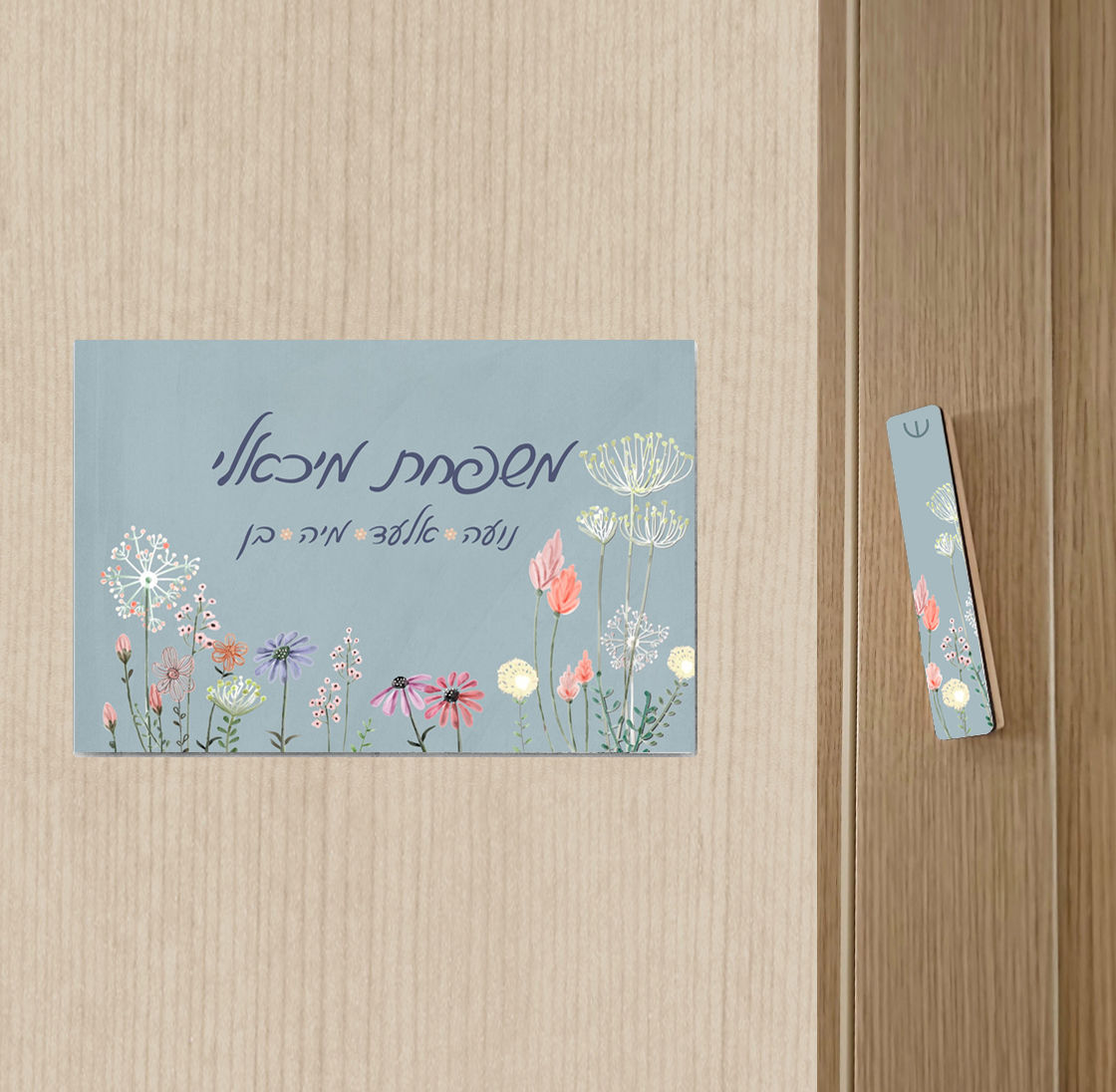 שלט לדלת בגוון כחול-אפרפר עם איורי פרחים עדינים צבעוניים בתחתית, לצד מזוזה תואמת בעיצוב זהה, תלויים על דלת עץ בהירה.
