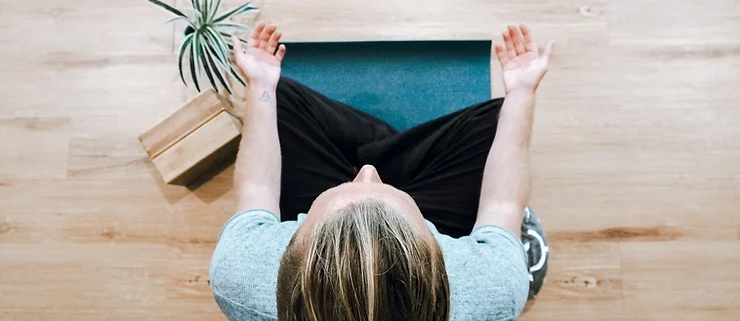 Person meditating on a blue mat, sitting cross-legged with palms up. Wooden floor, plant, and blocks in the background. Calm atmosphere.
