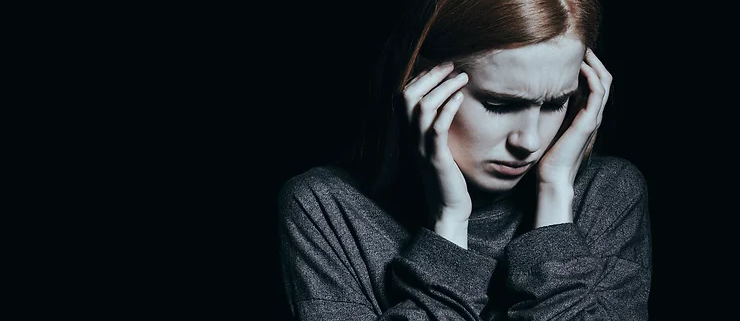 Woman in gray sweater holds her head in distress against a dark background, conveying stress or worry. Brown hair contrasts with the black setting.
