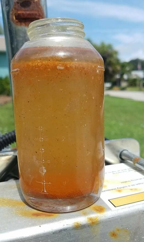 Clear jar filled with muddy orange liquid sits on a metal surface outdoors, with grass and blue sky blurred in the background.