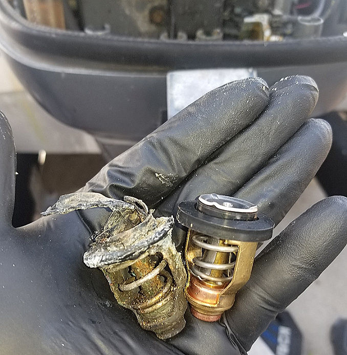 Gloved hand holding a heavily corroded and damaged old thermostat next to a cleaner replacement thermostat.