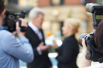 Photographers focus cameras on a man and woman engaged in conversation outdoors, blurred in the background. Urban setting and professional mood.