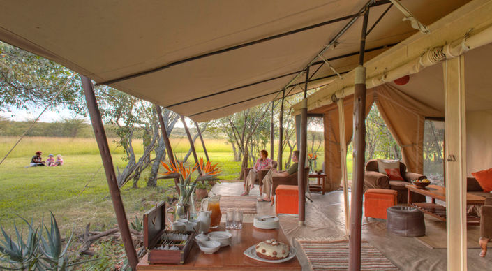 Table and chairs overlooking the Naboisho safari plains with a family sitting on the grass in the distance at Encounter Mara in Naboisho with Safari Ambitions