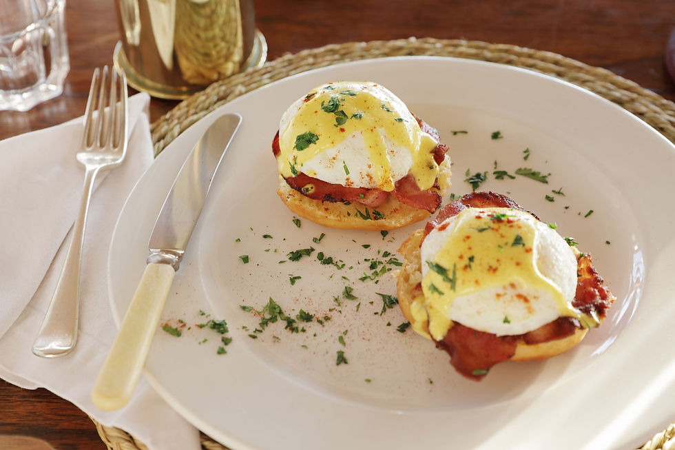 Two eggs Benedict with hollandaise and herbs on a white plate, with knife and fork on a napkin at a wooden table.