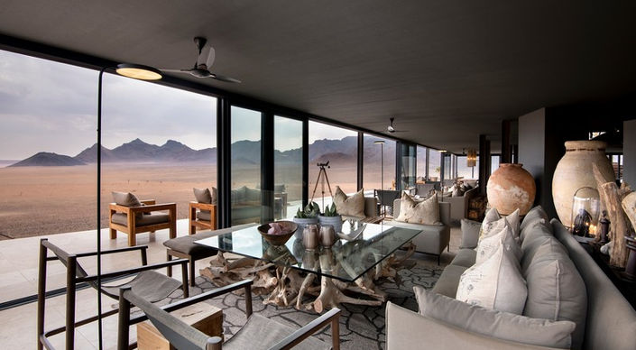 Large sofas and armchairs in the guest drawing room with glass windows an doors leading to a deck overlooking the desert at Sossosvlei Desert Lodge in Sossusvlei in Namibia