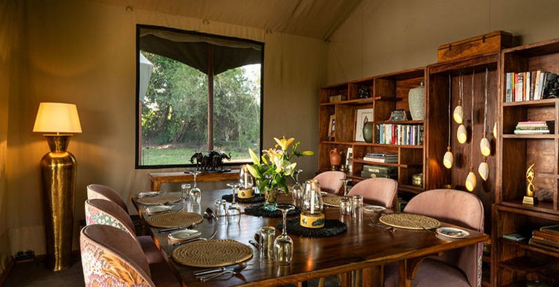 Laid dinner table with 6 pink armchairs in the main mess tent with bookshelves in the background at Rekero Safari Camp in The Masai Mara with Safari Ambitions. See the Great Migration here