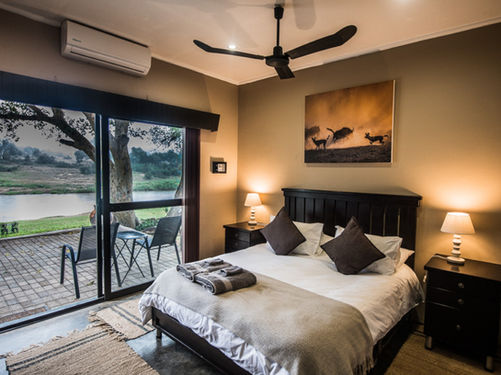 Double bed with beside lamps and central fan leading to glass doors to a deck with two chairs overlooking Olifants River