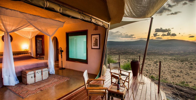 Open hillside guest bedroom with double bed lit up and a deck with chairs overlooking the view at Saruni Samburu Safari Lodge in Samburu, Kenya