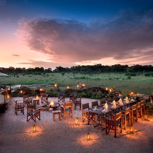Sunset at Somalisa Camp with camp chairs and a laid table with lanterns and a camp fire