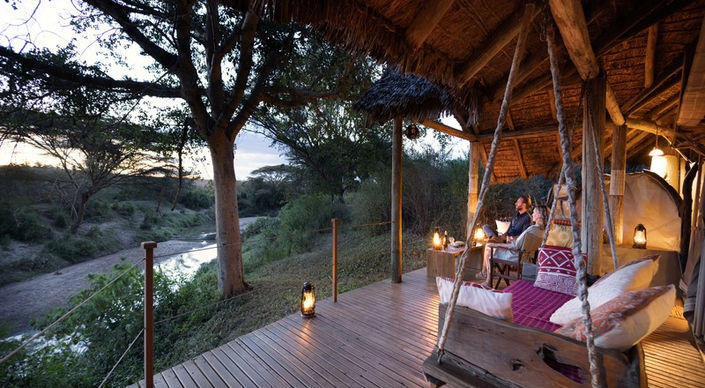 Deck lit up with lanterns and a swing chair with cushions at Basecamp Masai Mara Camp by the Talek River in The Masai Mara in Kenya